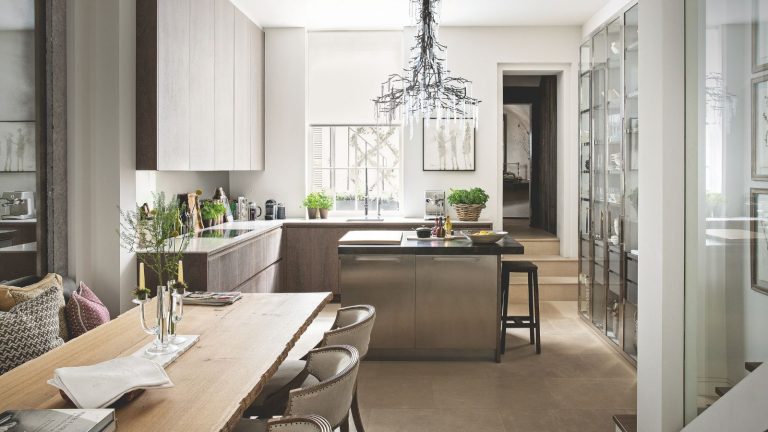 7 façons sournoises de cacher les courants d’air dans votre maison – et comment les corriger pour que les invités en vacances se sentent à l’aise et bienvenus A neutral kitchen with tiled flooring and white walls. In the center is an island with stool seating, and around it are wooden cabinets and white countertops. To the right are glass-fronted cabinets, and in the foreground is a wooden dining table with white chairs around it.