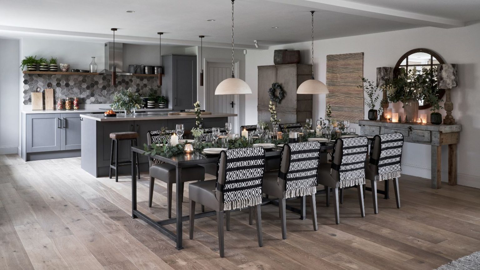 A black, white and grey kitchen with grey cabinetry, and wooden floors. There's a grey kitchen island in the background, and a large laid dining table in front, covered in branches, glasses, lit candles, with black and white blankets on the chairs. To the right is a circular mirror. The walls are white.
