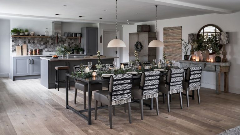 6 parfums d’automne que j’achète chez Net-a-Porter – Élevez votre maison et créez l’ambiance pour les fêtes de fin d’année cette saison A black, white and grey kitchen with grey cabinetry, and wooden floors. There's a grey kitchen island in the background, and a large laid dining table in front, covered in branches, glasses, lit candles, with black and white blankets on the chairs. To the right is a circular mirror. The walls are white.