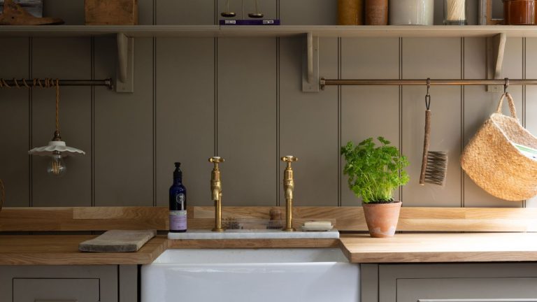 5 erreurs de lessive d’automne qui font que tout, du linge de lit aux vêtements, sentent l’humidité – et que faire à ce sujet A farmers sink with gold faucets, set into a wooden countertop. Greige wall panelling behind, leading to matching shelving.