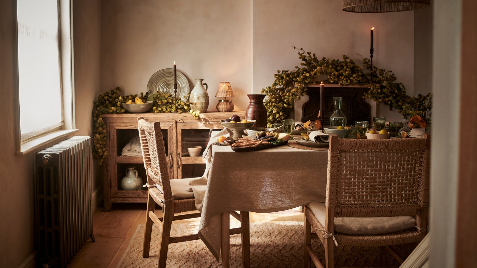 Une salle à manger rustique avec une guirlande d'automne sur la cheminée. Une table rectangulaire drapée d'une nappe en lin crème et des chaises en bois s'y approchaient. Une vitrine en arrière-plan avec de la vaisselle à l'intérieur. 