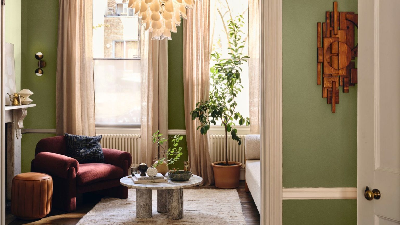 Comment les tapis affectent réellement les courants d’air – et la manière la plus intelligente selon les professionnels de garder votre maison au chaud en novembre A cozy apartment living room with green painted walls. A red armchair in the far corner, a small round marble coffee table in front of it, and a cream rug.