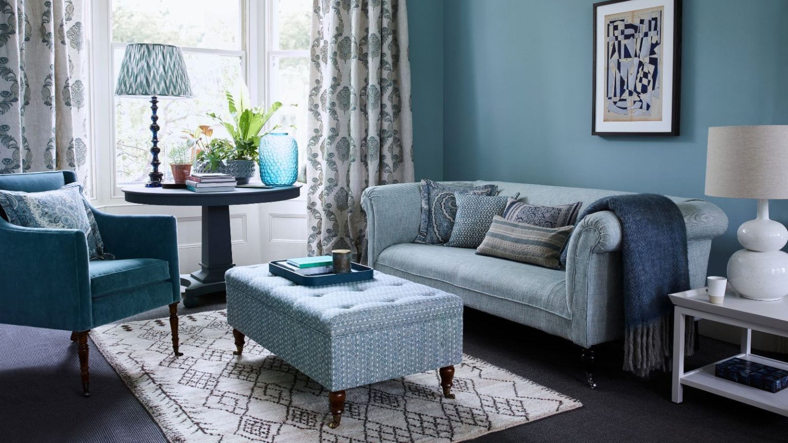 « C’est la façon naturelle de signaler le calme » – Le parfum du soir est une nouvelle tendance automnale relaxante que j’adopte pleinement cet automne A blue living room with brown wooden floors, and large bay windows with patterned curtains. There's a blue couch, blue upholstered chair and blue patterned storage ottoman, on top of a patterned rug, and a circular table under the window with a lamp, vase and house plant. To the right of the sofa is a dark blue throw, white table and white ceramic lamp with white shade.