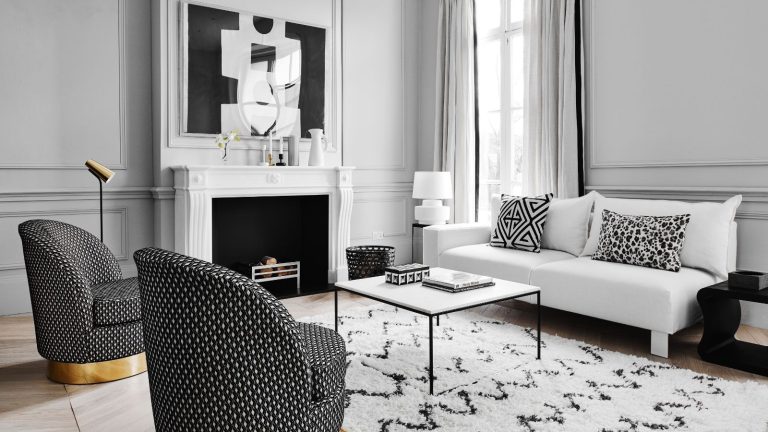 A black and white living room with white walls, a white mantelpiece, and white curtains, and a black and white artwork above the fireplace, a black and white rug, a white couch with black and white cushions, a black and white coffee table and two black and white chairs with gold bases.