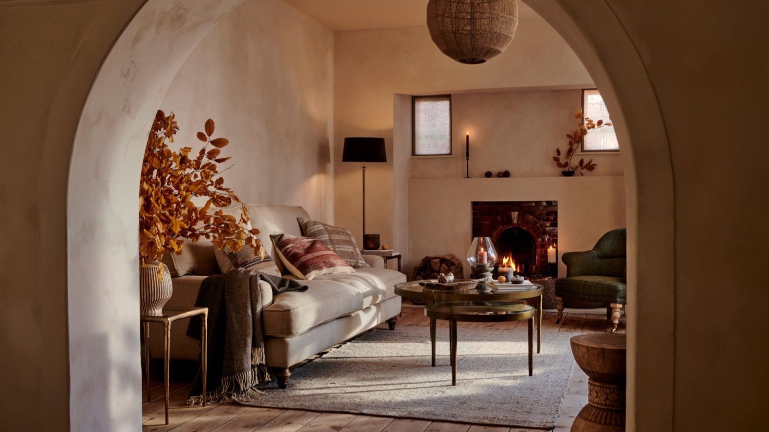 4 façons non toxiques et zéro déchet d’une artiste olfactive et parfumeur de parfumer sa maison pour l’automne – des traditions danoises et des effets gourmands, à l’introduction de la nature à l’intérieur A cozy fall-esque living room viewed through an archway. A cozy cream sofa with green and red throw cushions behind a round coffee table. A large orange foliage arrangement in a vase beside it.
