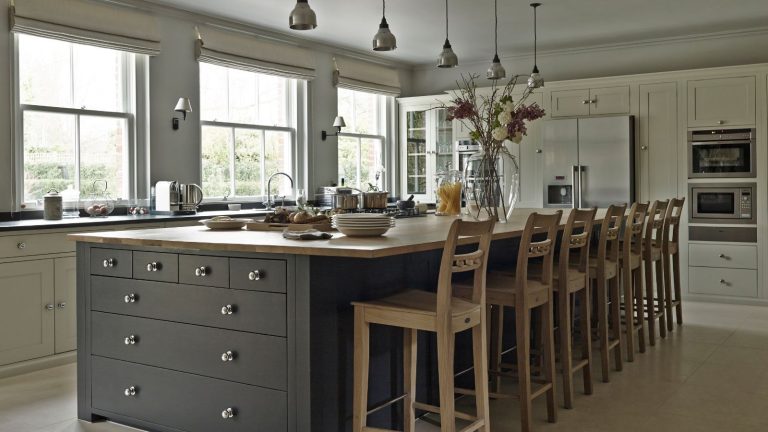 Arrêtez les odeurs de poubelles de janvier avec cette astuce facile au citron surgelé. Les professionnels du nettoyage ne jurent que par A large modern kitchen with tiled flooring, white cabinets, and a large kitchen island with grey drawers and a wooden countertop. Behind the island are seven tall wooden chairs facing large windows.