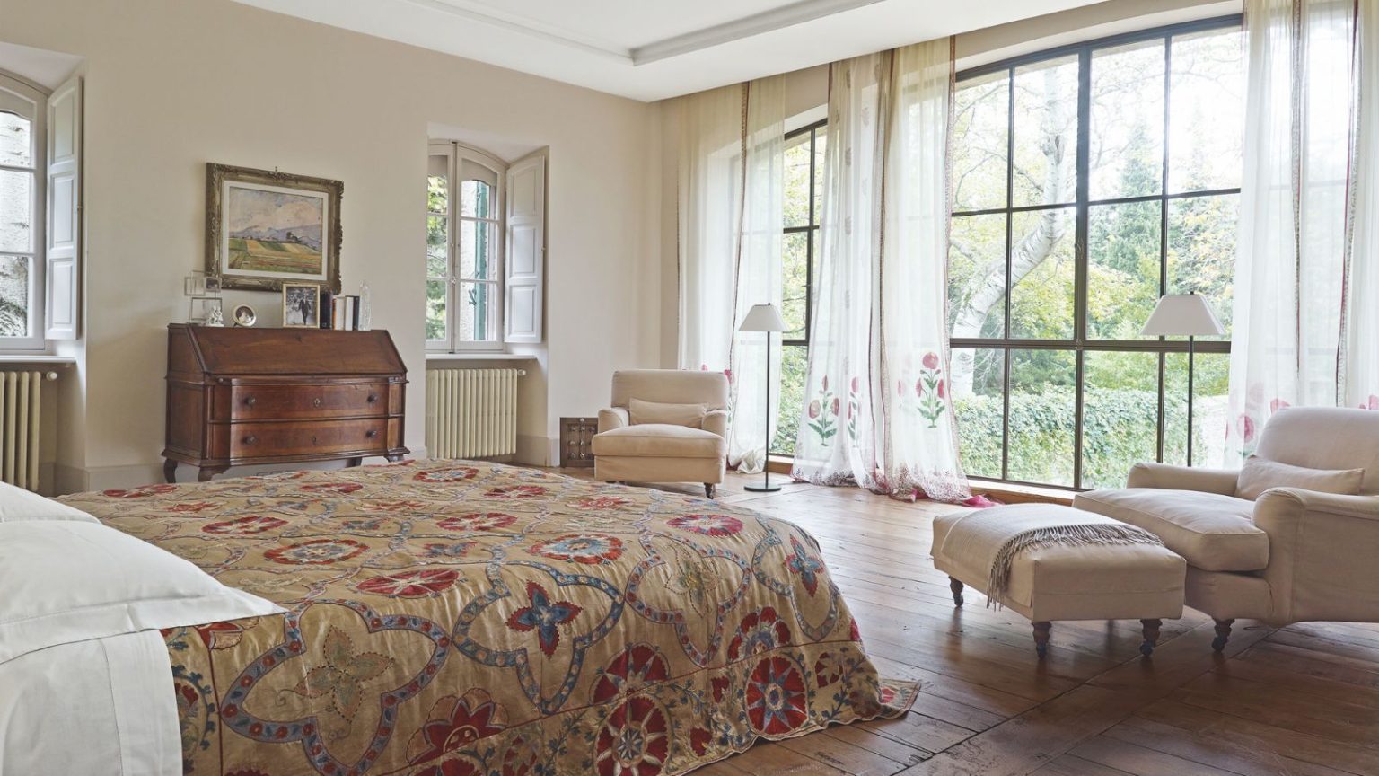 Pulvériser ce spray d’huile essentielle facile à faire soi-même autour de vos fenêtres en octobre dissuadera les araignées – elles détestent l’odeur. A bright bedroom with large windows with sheer curtains, brown wooden floors, a large bed with a patterned quilt, matching cream upholstered chairs, and a wooden chest of drawers against the opposite wall.