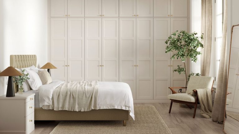 A neutral bedroom with large built in closets along the back wall with panelled walls. A double bed in front of them with two symmetrical night stands, a chair opposite