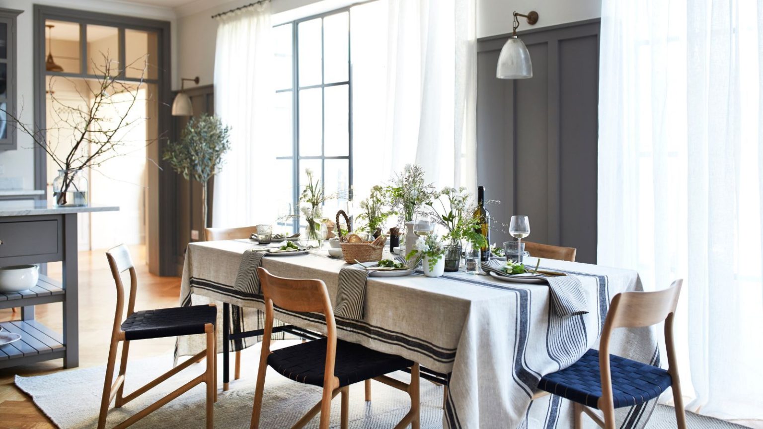 Les designers disent que c’est la formule parfaite pour éclairer une salle à manger afin de la rendre confortable et propice à la conversation – et cela ne signifie pas abandonner l’éclairage zénithal. A bright and airy dining room with a striped tablecloth on a long table, set with wine glasses, wine, and napkins, and blue and brown chairs on top of a white rug. Behind are brown panelled walls, and sheer curtains over crittal windows.