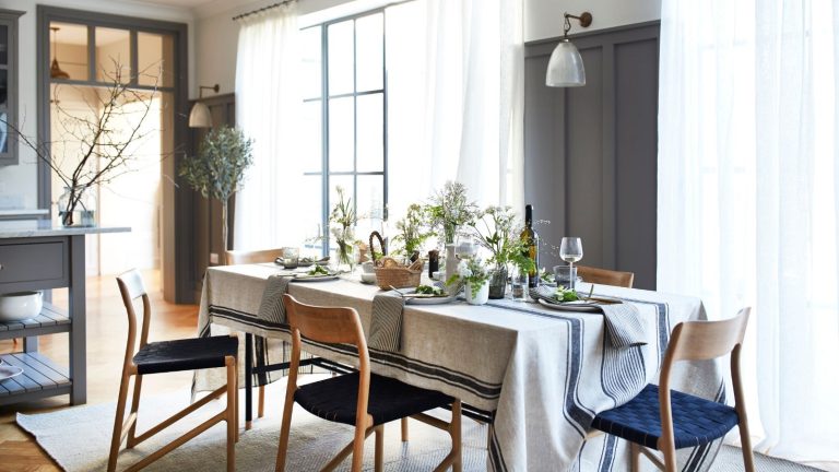 A bright and airy dining room with a striped tablecloth on a long table, set with wine glasses, wine, and napkins, and blue and brown chairs on top of a white rug. Behind are brown panelled walls, and sheer curtains over crittal windows.