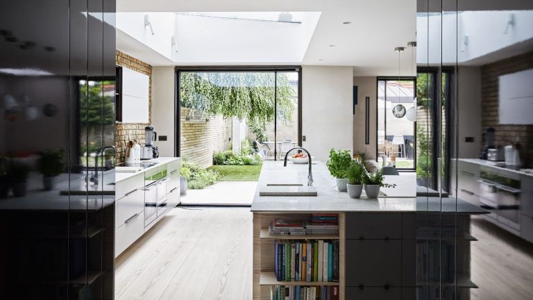 A bright and airy modern kitchen with wooden flooring, black and white cabinetry, an island with white marble countertops and built-in shelving filled with recipe books, an exposed brick backsplash, and large window doors to a lush green garden.