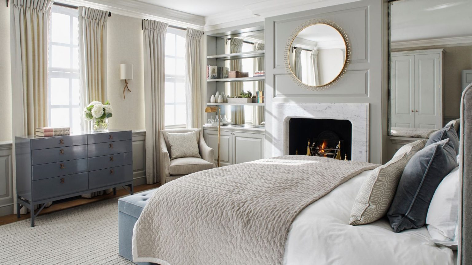 L’échange de garde-robe d’automne – 5 conseils de pro pour rationaliser et ranger votre garde-robe pour les mois les plus froids A neutral bedroom with large double bed, cream rug, blue chest of drawers, glass-backed shelves, a round mirror, white closet, and a white marble fireplace with a fire burning.