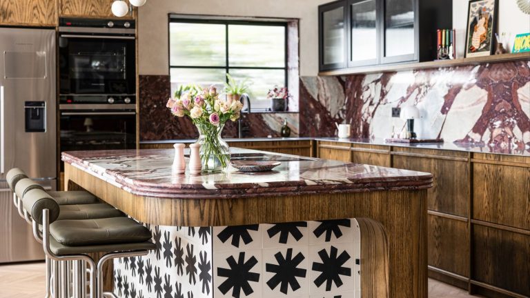 A kitchen with rustic wooden cabinets, and red marble counters and backsplashes. A matching island with black and while tile surround.