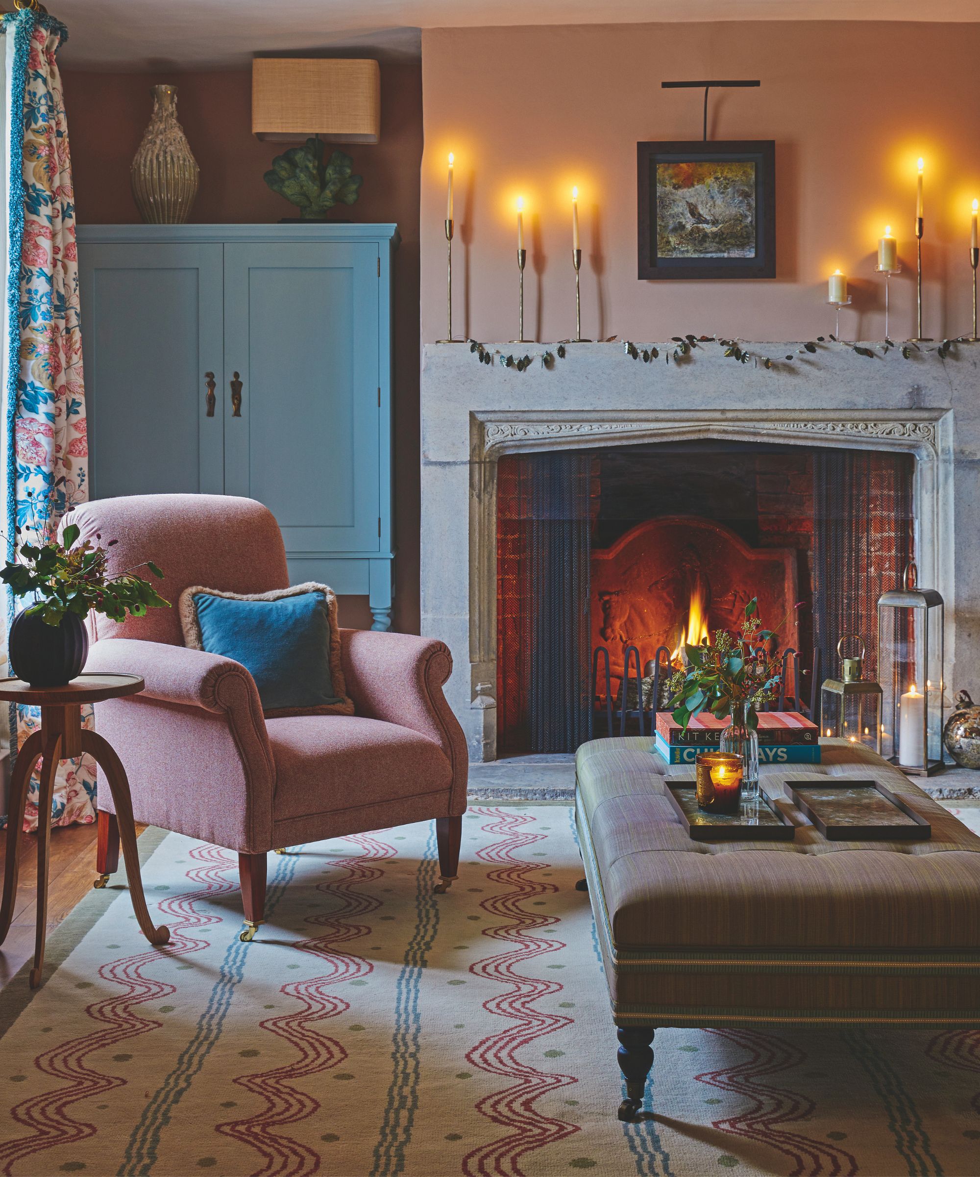 Un salon confortable rose et bleu avec un feu allumé dans la grande cheminée en pierre, des bougies, un tapis à motifs, une chaise rose, une table d'appoint en bois, un meuble bleu et un grand pouf avec une bougie et un vase de fleurs.