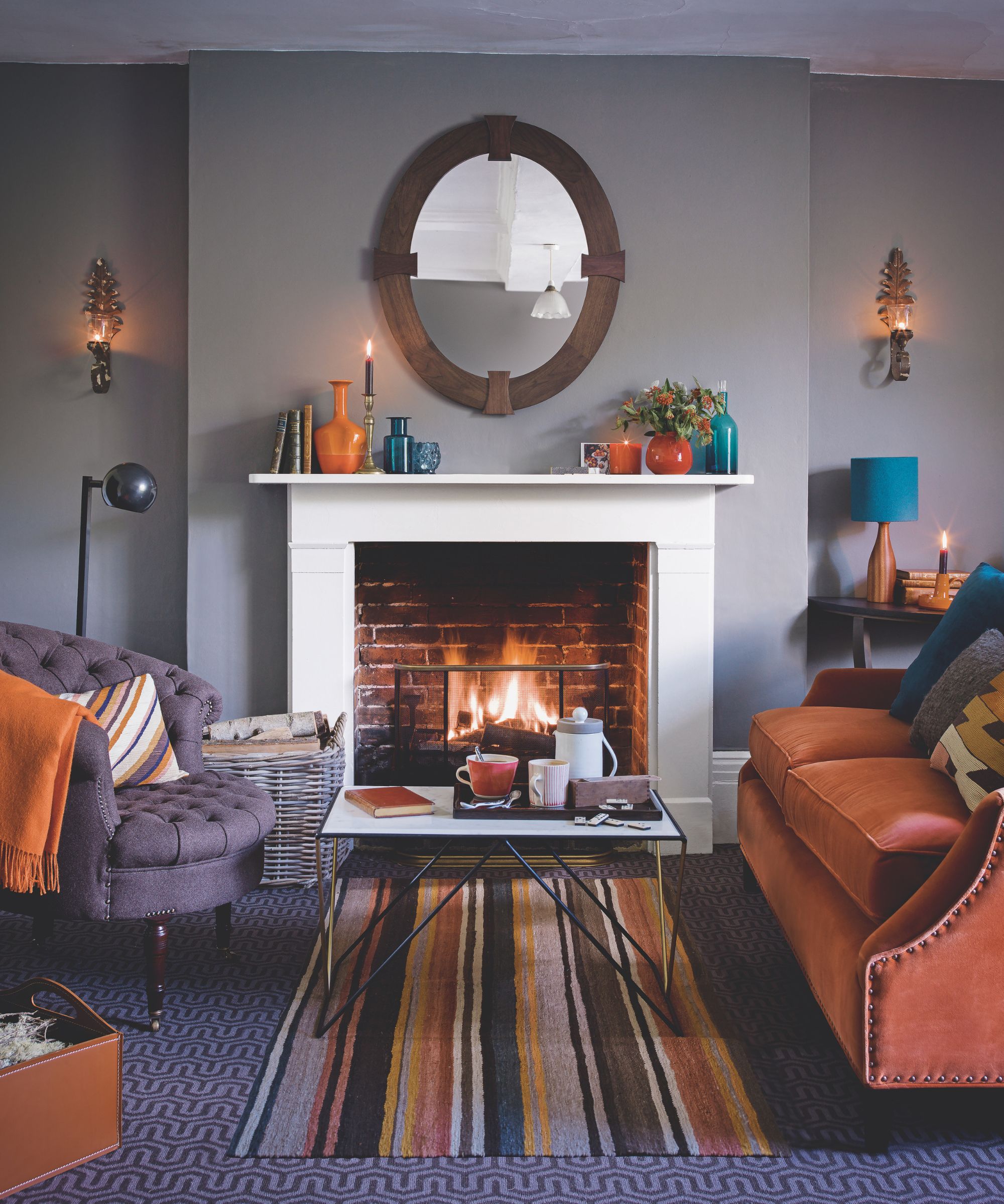 Un salon lilas avec une cheminée blanche, des chaises et des canapés orange et violets avec des coussins assortis et un tapis rayé sous une table basse au centre de la pièce.