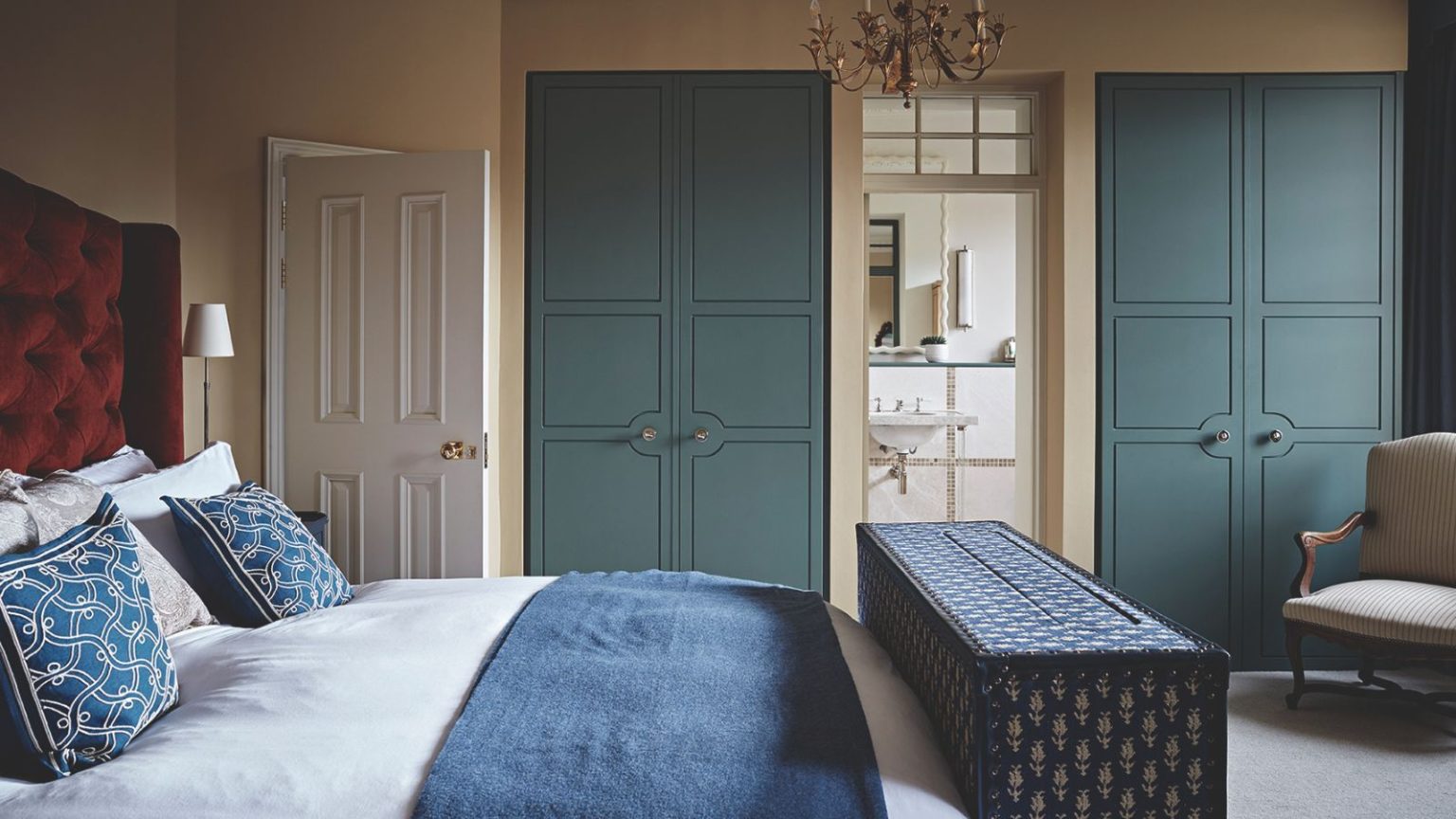 Les créateurs d’une seule couleur évitent toujours dans les chambres d’amis (et quoi utiliser à la place) A bedroom with blue built-in closets, a red velvet headboard, white bedding, blue cushions, and a blue storage ottoman at the end of the bed.