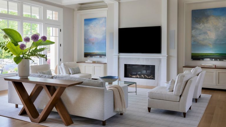 White living room with tall ceilings, fireplace with mounted TV above, hardfloors and rug, with sofas set to center the TV. Big windows give lots of natural light and a wooden console in the foreground showcased enormous purple blooms and large green leaves