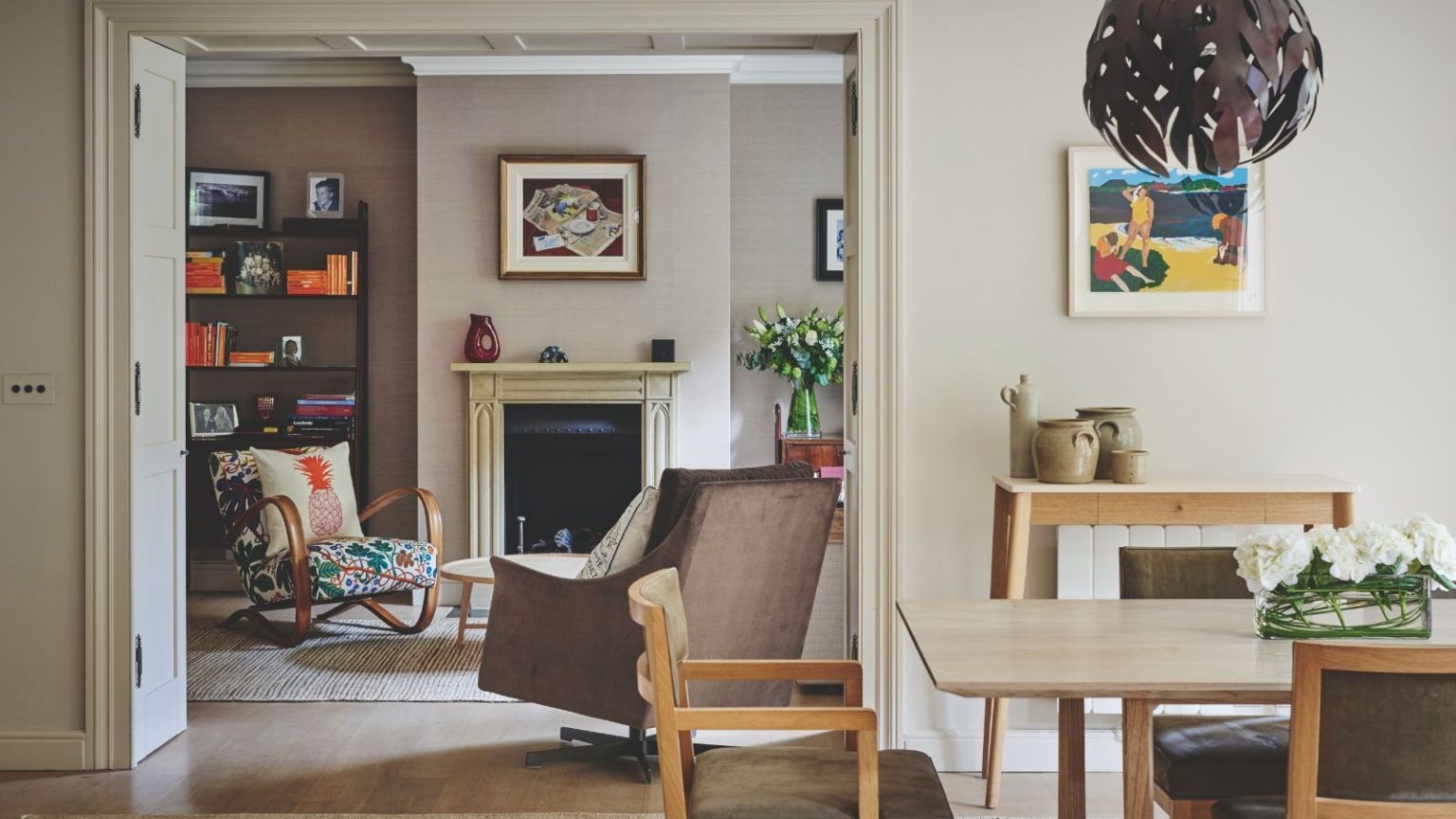 La « différence entre la vie et la mort » : des experts en entretien ménager révèlent 3 tests de détecteur de fumée que vous devez effectuer en octobre A neutral dining area with wooden chairs and table, and a doorway through to an eclectic neutral living room with upholstered chairs, a black fireplace with cream mantelpiece, artwork, flowers, and a black shelving unit filled with books.
