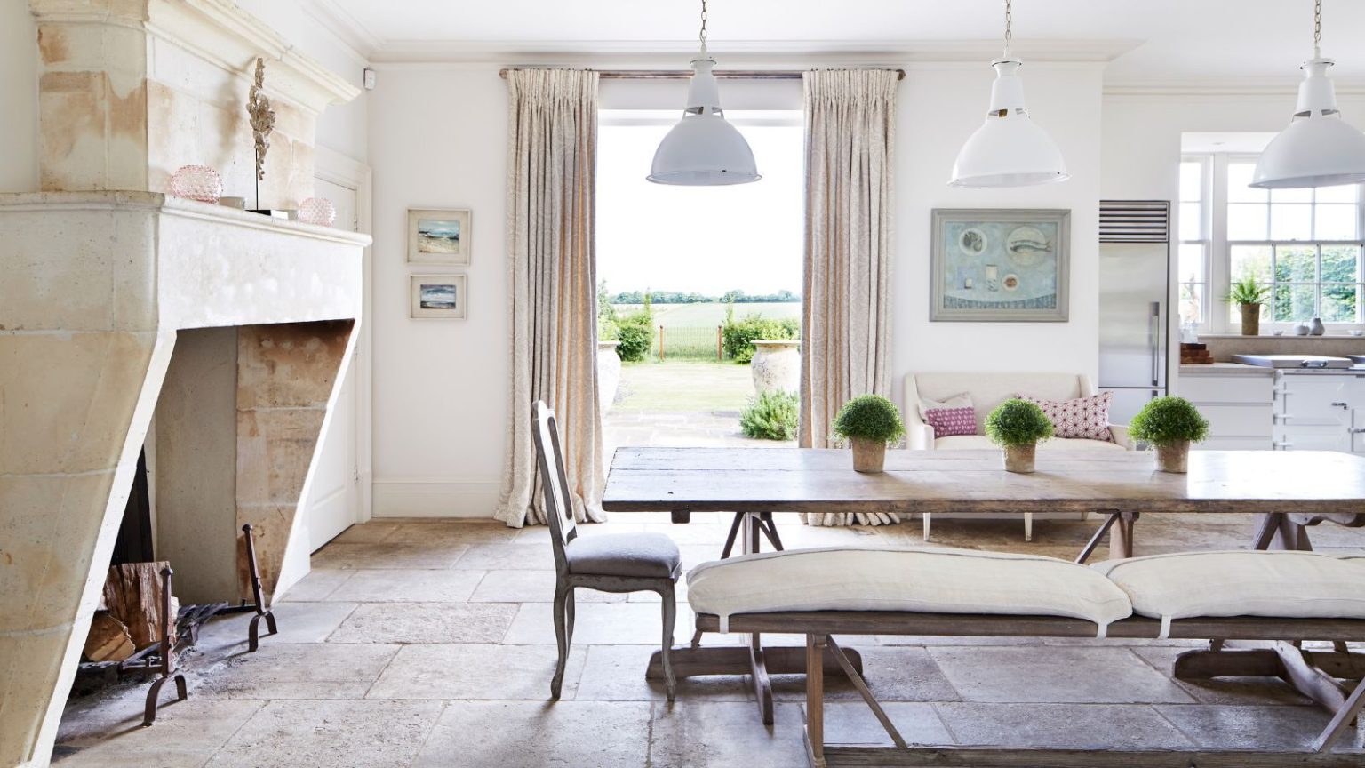 « Lorsque votre espace est bien rangé et que vos essentiels sont disposés, vous commencez votre journée avec clarté, sans encombrement » – Ma réinitialisation du soir de 10 minutes est la clé pour des matinées sans stress A large kitchen diner with a large dining table with cushioned wooden benches, flagstone floor and a huge stone fireplace feature to the left. Behind is a white AGA, and open doors with neutral curtains out to a large garden. Above are three large white lamps.