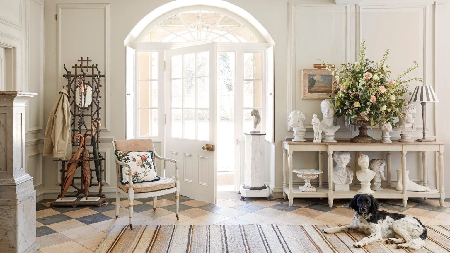Je prédis une vente à guichets fermés ! Avec quatre chiens à fourrure à la maison, ce Miele est le seul aspirateur par lequel je ne jure que – et il est maintenant réduit de 209 $ Bright and airy entryway with a striped rug on tiled patterned floor, a black and white spaniel, open glass-panelled white door, wooden umbrella stand and wooden sideboard covered in busts and flowers. The walls are white and panelled.