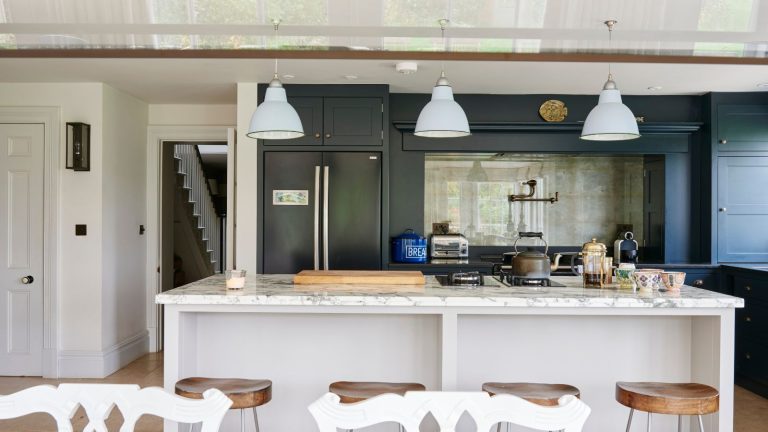 A bright and airy kitchen with a white marble island, four wooden barstools, dark green cabinets and three hanging white pendant lights