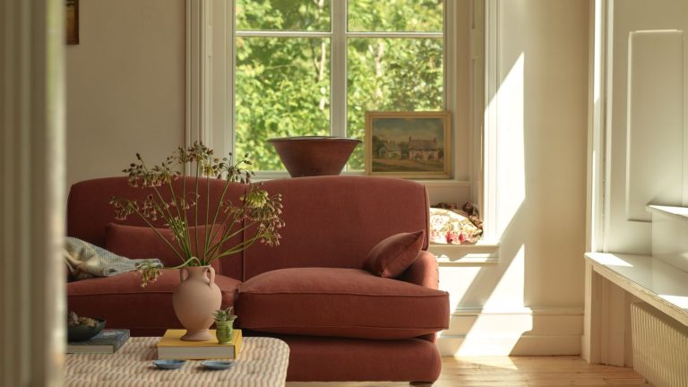J’ai essayé 3 principes du Vastu Shastra, le « yoga du design », et cela a rapidement calmé et équilibré ma maison encombrée – cela a également fait disparaître le stress et l’anxiété. A red traditional sofa in a white living room with large windows. Behind an upholstered ottoman table with a vase of large flowers on it.