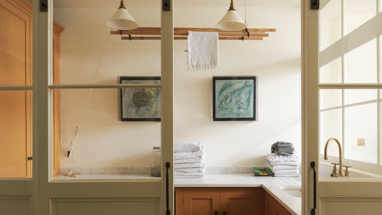 « Tout simplement incroyable, cela fonctionne vraiment » : des milliers d’acheteurs Amazon ravis ne jurent que par les pouvoirs anti-odeurs des Oxi Odor Blasters pour les vêtements sales. Wooden and cream laundry room with hanging dryer and glass sliding doors. The space has lower cabinets only with a mid-century wooden finish, white marble counters with short upstand, and there are folded towels visible.