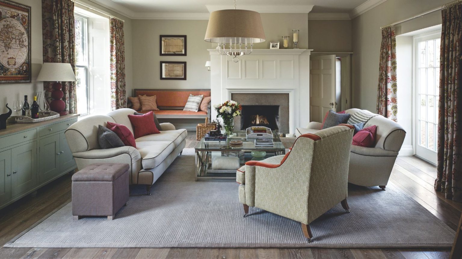 7 choses que je dis à chaque propriétaire sur l’entretien de la chaudière en tant que professionnel du CVC pour éviter une panne lors de la nuit la plus froide de l’année A cozy living room with large rug, upholstered chairs and matching sofas, a glass coffee table, and a large white panelled fireplace. To the left is a blue sideboard. There are bright windows either side with floral curtains.