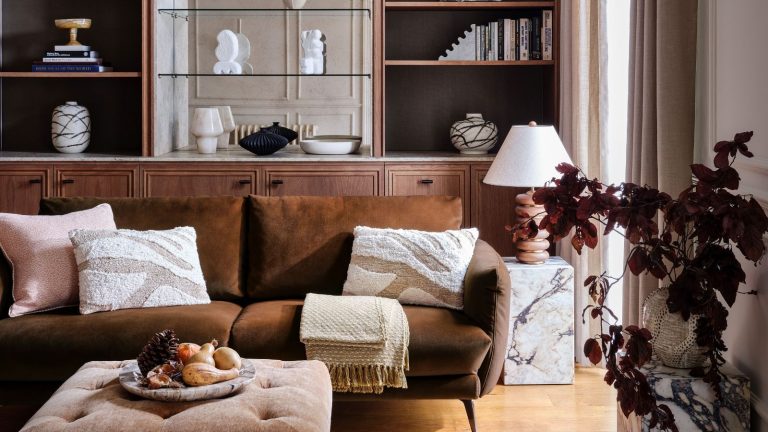 3 choses que j’organise en octobre 2025 pour ramener le calme à la maison avant que la haute saison des accueils ne batte son plein A brown velvet sofa in a neutral living room. Large wooden built-in shelves behind, a small marble table beside it, and a brown-leaf plant in the foreground.