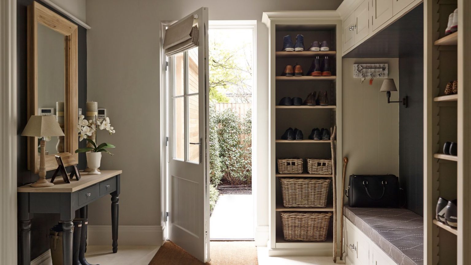 Le montage très britannique : les premières impressions comptent – Pourquoi l’entrée printanière prépare le terrain pour tout ce qui suit A bright entryway with an open glass-frame door, black side table with mirror above to the left, with lamps and flowers on it, and shelves and a bench to the right.
