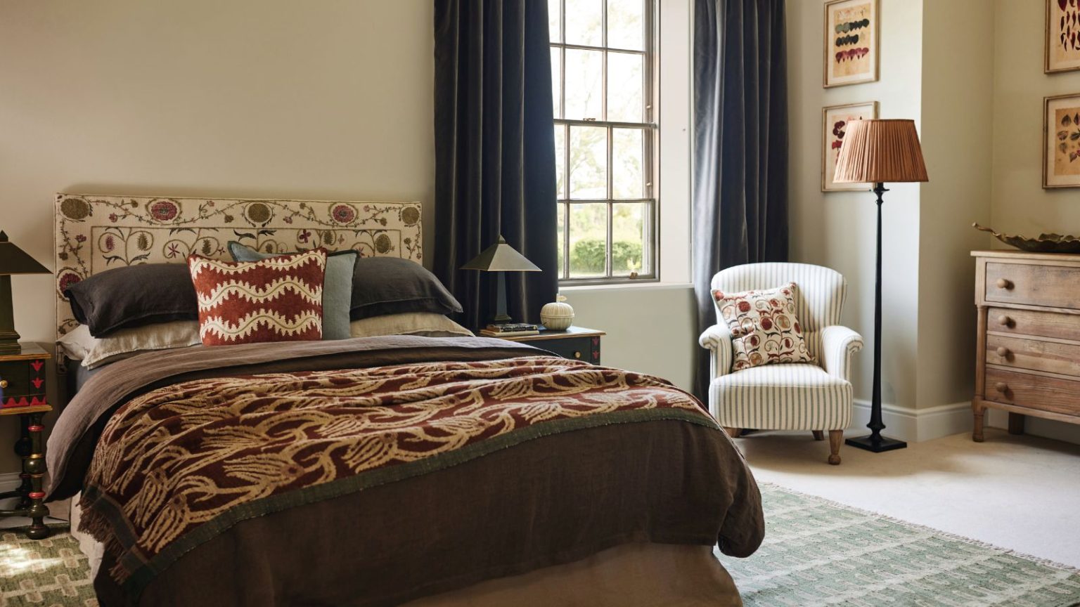 Ne transportez pas d’allergènes d’été jusqu’à l’automne : c’est le seul article ménager que vous devez laver à la fin de l’été pour éviter les nez bouchés et les démangeaisons. An autumnal bedroom with brown bedding, a patterned headboard, and a white chair beneath a lamp in the corner beside a window.