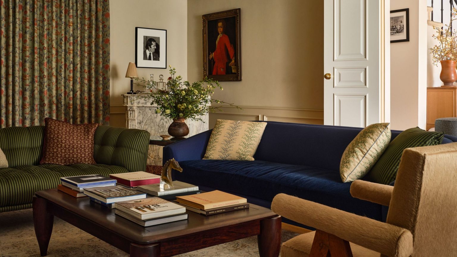 Un renouveau colonial des années 1940 où les couleurs et les motifs se mélangent avec l’ancien et le nouveau pour créer une maison familiale véritablement transitionnelle Cozy living room with a navy velvet sofa, a striped olive green sofa, an orange upholstered armchair, and a dark wood coffee table in front of a marble fireplace
