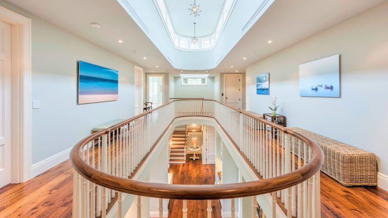Dilemme de stockage : « Je ne supporte pas les piles d’escaliers car personne ne les monte jamais à l’étage » – cette solution à 50 $ fonctionne avec le problème, pas contre lui. Grand hallway with coastal accents such as wicker benches and seaside art on walls. The stairs are in view in the background, with an oval hallway railing and gallery view over the lower floor. The floors are hardwood and the ceiling is brigh with a matching oval window gallery