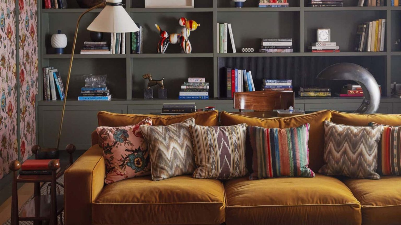 J’ai du mal à me concentrer lorsque je fais du rangement, mais la méthode des pistes de ski me facilite la tâche, même avec un programme hivernal chargé – c’est parfait pour les esprits neurodivergents A maximalist living room with a mustard velvet sofa in front of built-in grey book shelves.