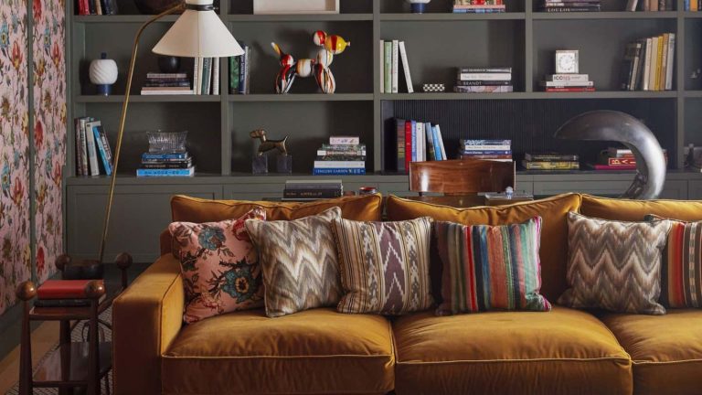 J’ai du mal à me concentrer lorsque je fais du rangement, mais la méthode des pistes de ski me facilite la tâche, même avec un programme hivernal chargé – c’est parfait pour les esprits neurodivergents A maximalist living room with a mustard velvet sofa in front of built-in grey book shelves.