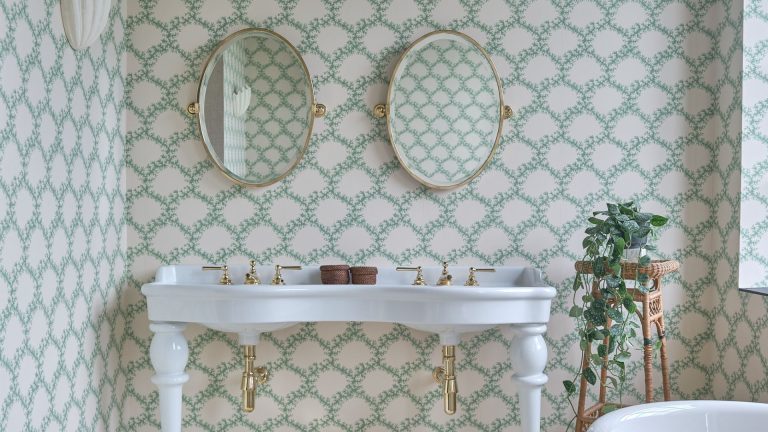 Vous en avez assez de la moisissure dans votre petite salle de bain ? En tant que spécialiste de la qualité de l’air, c’est la seule chose que vous faites de mal et la solution pour éviter un hiver misérable et moisi. A his and her bathroom sink with two ovel mirrors above each sink. There is pretty green and white wallpaper on all of the walls and a rattan stand with a trailing plant sitting on top.