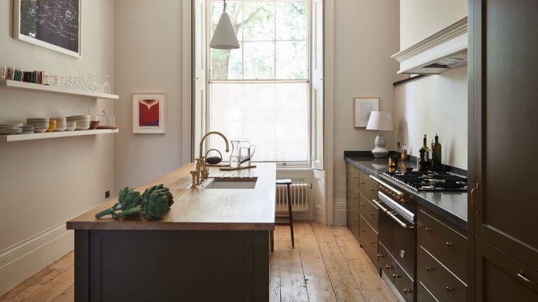 Ce génie Joseph Joseph Caddy à 39 $ a instantanément vidé mon comptoir bondé – L’hébergement de vacances dans ma petite cuisine est devenu tellement plus facile Compact cream galley kitchen with black wooden cabinets and wood-top island and floating wall shelves. There is a large window at the end and a vintage radiator just in view