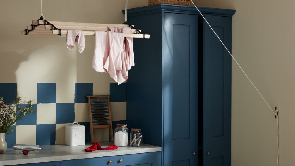C’est le seul article que vous lavez trop cet hiver – Les experts en lessive révèlent la fréquence réelle nécessaire pour protéger cet essentiel par temps froid. Dark blue painted cabinets with matching blue and white tiles in a small laundry room. A ceiling-suspended drying rack above, with some pink clothes on it.