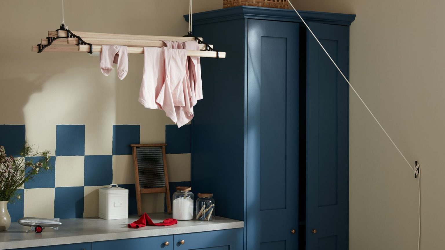 Les professionnels de la blanchisserie disent qu’il est temps de mettre fin à cette règle obsolète de la laveuse : vous économiserez du temps et de l’argent avec ce simple changement Dark blue painted cabinets with matching blue and white tiles in a small laundry room. A ceiling-suspended drying rack above, with some pink clothes on it.