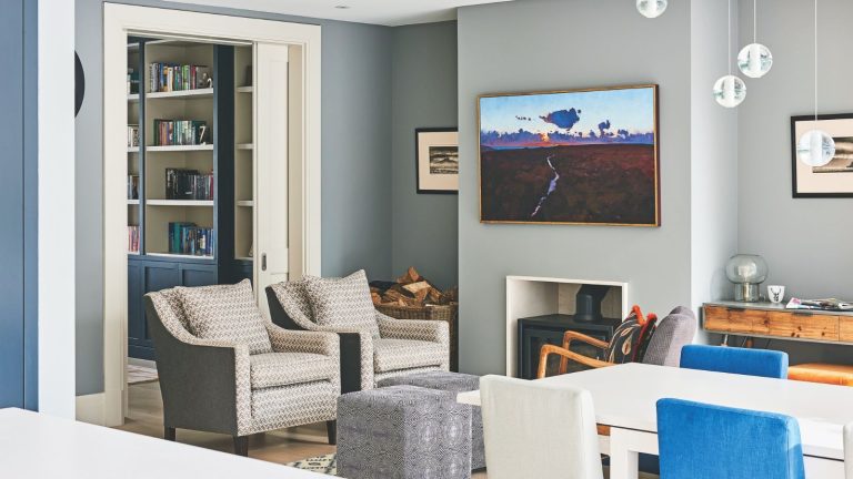 Les experts en CVC révèlent exactement le nombre de déshumidificateurs dont vous avez besoin pour lutter contre la condensation et la moisissure cet hiver A grey living room space, with a central black log burner with a large artwork above, two matching patterned upholstered chairs, two matching cubic ottomans, and a grey upholstered chair with wooden handles and a patterned cushion. To the right foreground is a white dining table with blue and white chairs. To the left, a room is seen behind the matching upholstered chairs, with blue and white bookshelves.