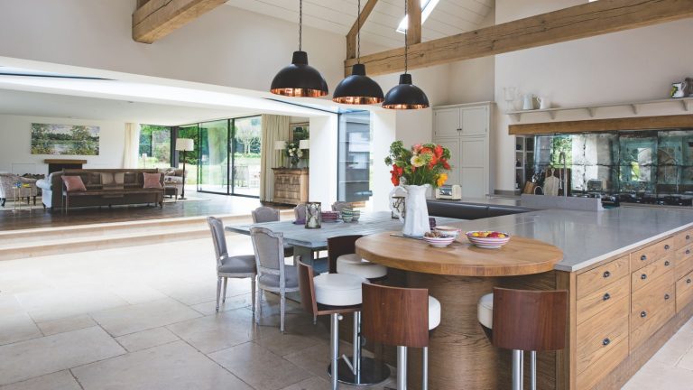 Ce produit de remplissage en laine d’acier à 22 $ élimine efficacement les souris de vos murs – les acheteurs disent que c’est « tout ce dont vous avez besoin dans une seule boîte » A spacious kitchen with tiled flooring and exposed wood beams. In the foreground is a large brown cabinet island with silver countertops, a circular wooden table, and a white dining table and chairs. On the island is a vase of flowers. In the background is a living space with a brown leather sofa, and large window doors out onto a green garden.