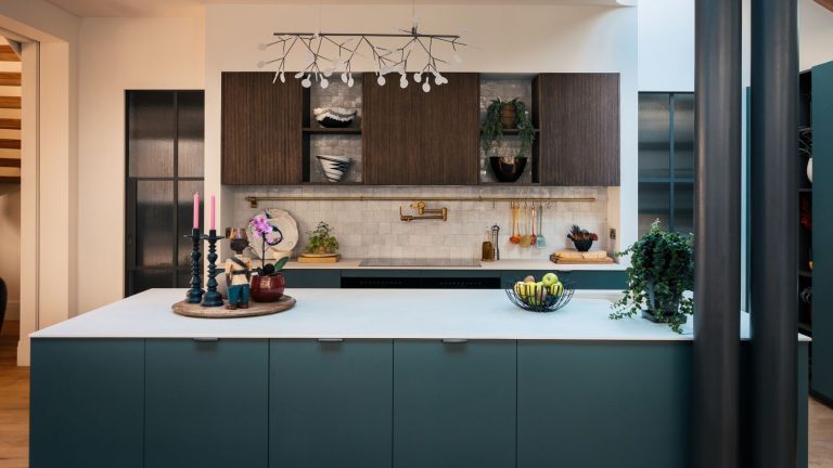 Ces étagères empilables à 15 $ changent totalement la donne pour l’organisation de la cuisine avant les fêtes de fin d’année chargées. A blue kitchen with large island with white countertop, stove top with white Zellige tiled backsplash with wooden cabinets above, a statement lighting fixture, and two exposed beams to the right.
