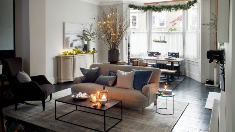 Cette bougie Diptyque réduite est la préférée de Victoria Beckham et elle est parfaite pour mettre les invités dans l’ambiance des vacances avec ses notes de baies. A cozy festive living room with a large cream sofa, coffee table with lit candles, large rug, brown upholstered chair, and a dining table under large bay windows with white sheer curtains. In the centre of the room is a large vase with lit dry twigs, and a tall candlestick. Above the window, festive greenery is hanging, and to the right is a white side-table with fairy lights, a vase of festive sprigs, and an artwork above.