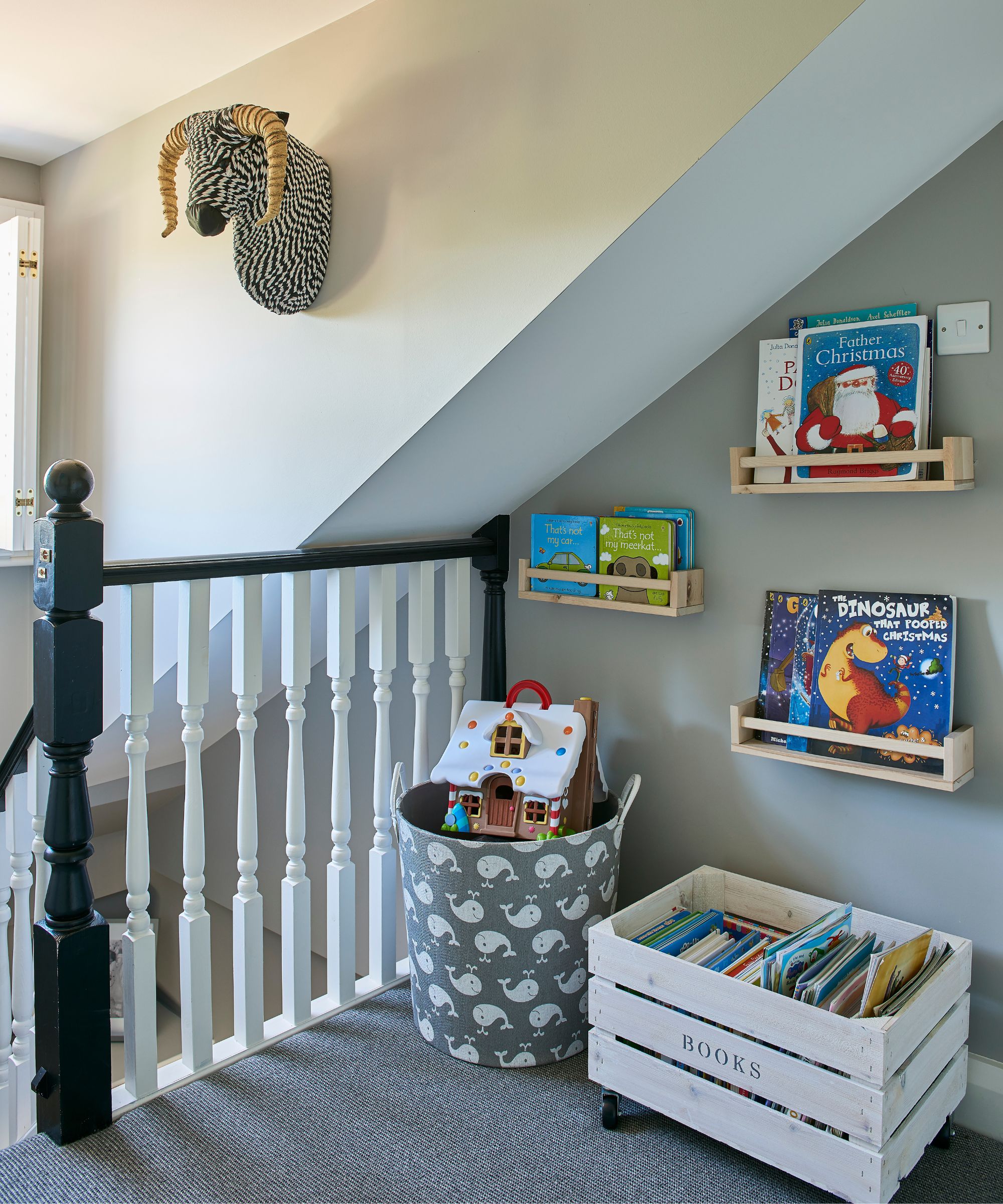 Un espace soigné et bien rangé en haut d'un escalier noir avec des rampes blanches avec des livres pour enfants organisés sur des étagères, et dans une caisse en bois blanche au sol à côté d'un panier de rangement rempli de sols et imprimé de baleines. Le sol est recouvert d'une moquette gris foncé et les murs sont gris pâle.