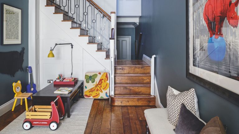 « Le secret d’un matin de Noël sans stress est de faire de la place maintenant » – Essayez cette astuce de désencombrement qui fonctionne réellement avec les enfants d’ici le 25 novembre A space with dark blue walls, a white staircase, and dark wood flooring, with a children's playroom area to the left with toys and a low, dark-wood desk on a white rug. To the right is an upholstered bench with mismatched cushions, and a large artwork of a red rhino standing on a blue ball above. The staircase is seen in the background.