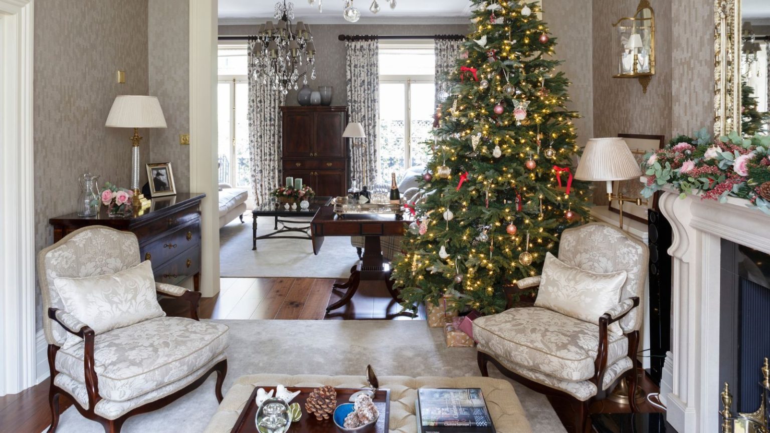 3 façons simples d’empêcher mon salon de ressembler à un désordre le jour de Noël – Anticipez le chaos et retrouvez le calme en quelques minutes A festive living room with a large lit Christmas tree to the right. In the foreground is a large square ottoman on top of a cream rug with matching cream upholstered chairs with wooden legs. To the right is a white mantelpiece with a floral garland. A room is seen beyond with a matching cream carpet and dark wood furniture, and two windows with white patterned curtains.