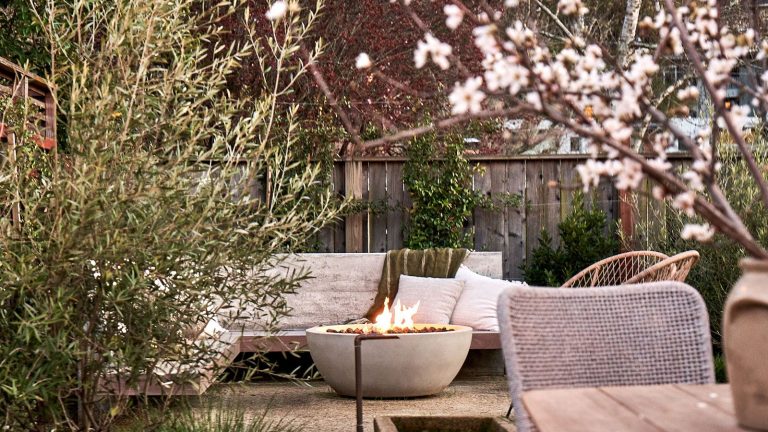 7 idées de conception recommandées par des experts pour de magnifiques jardins d’hiver dans des climats chauds – pour tirer le meilleur parti de vos choix de plantation dans des conditions plus douces fire pit with garden sofa, cushions and throws, shrubs and tree with blossom, and a table and chair