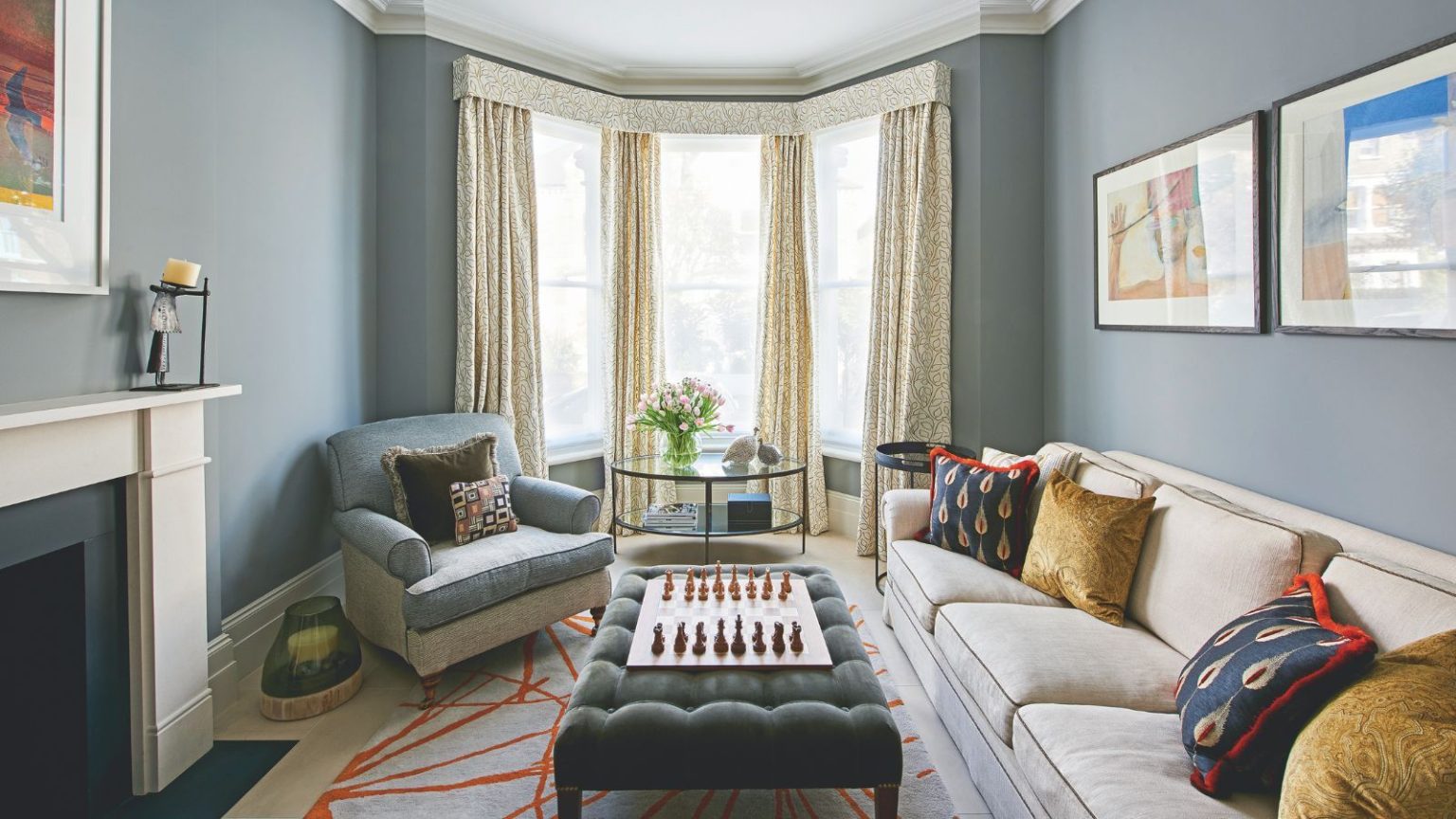 Ce purificateur d’air Dyson « indispensable » est compact, silencieux et élimine les odeurs en quelques minutes – il est parfait pour les petites maisons A blue living room with large bay windows with yellow curtains, a large cream couch with cushions, and a blue upholstered chair. In the centre of the room under the windows is a large circular glass table with pink tulips on it, and in the foreground is a green velvet ottoman on top of a patterned rug with a wooden chessboard on top. To the left is a white mantelpiece.