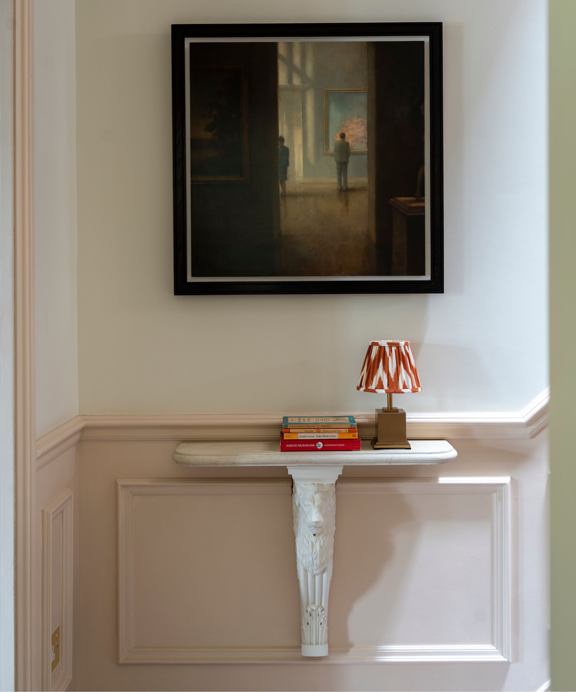 Un espace d'entrée avec des plinthes à panneaux roses, une table blanche fixée au mur, une pile de livres, une petite lampe avec un abat-jour rouge et blanc et une grande œuvre d'art au-dessus.