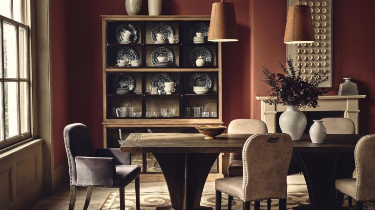 A deep red dining toom with a large wooden dining table. Upholstered chairs around it, and two hanging brown leather pendants above. A wooden cabinet of fine crockery in the background.